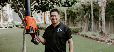 A smiling landscaping worker in a black shirt holding a red leaf blower outdoors.