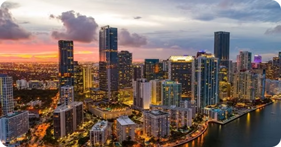 A skyline view of downtown Miami, Florida, along the Biscayne Bay waterfront at sunset, featuring illuminated high-rise modern skyscrapers and urban coastal architecture.