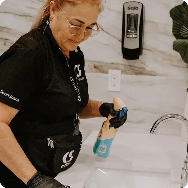 A CleanSpace commercial cleaning staff member wearing black gloves spraying disinfectant spray over a marble-backed sink.