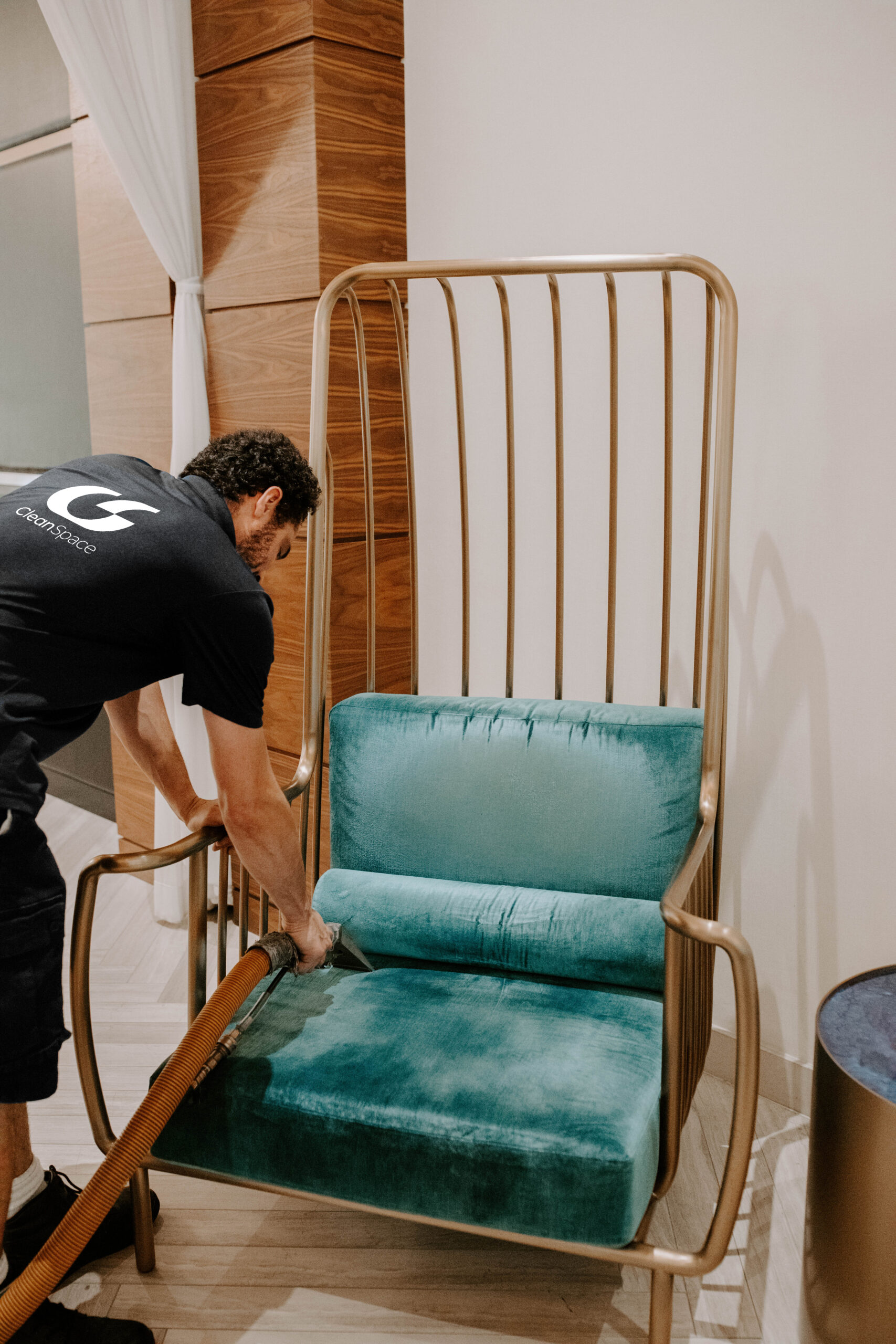 CleanSpace commercial cleaning technician using an upholstery extraction tool to deep clean a teal velvet armchair with a gold metallic frame.
