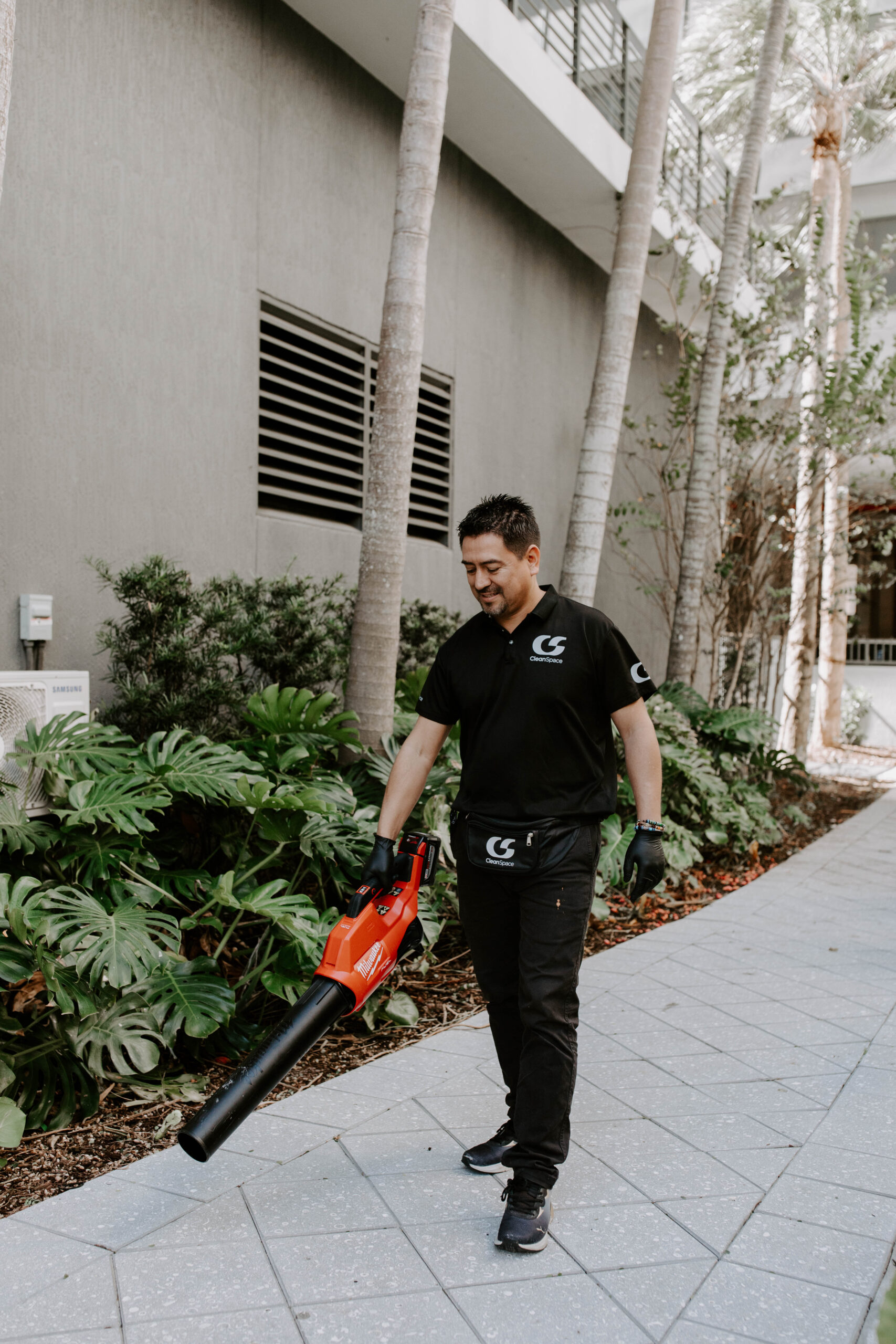 CleanSpace commercial cleaning technician using a red leaf blower on a tiled outdoor walkway along a building complex.