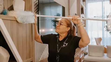 A female cleaner wearing glasses and a black uniform using a long duster to clean a wooden shelf.