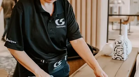 A professional cleaner wearing a branded CleanSpace polo shirt cleaning a wooden tabletop near decorative vases.