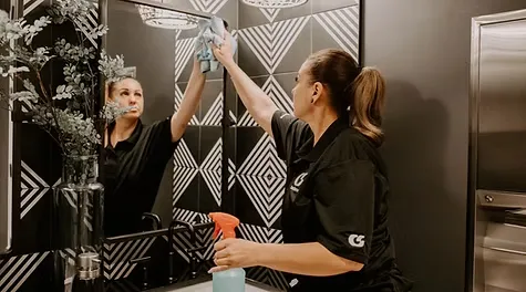 A female cleaner reaching up to wipe the top of a bathroom mirror with a cloth in a modern tiled bathroom.