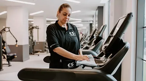 A female cleaner wiping down a treadmill console with a micro-fiber cloth in a modern gym.