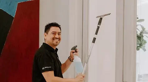 A smiling male cleaner holding a spray bottle and an extension squeegee pole to clean window panels.