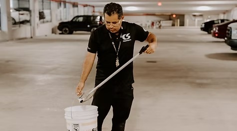 A male cleaner in a black uniform using a grabber tool to drop litter into a bucket inside a parking garage.