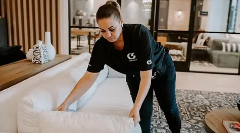 A female cleaner wearing a black CleanSpace uniform arranging white cushions on a modern sofa.