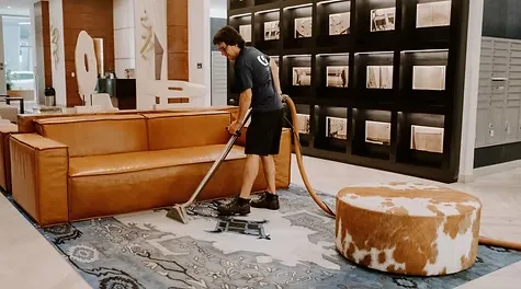 A male cleaner using a carpet extractor wand to clean a patterned rug next to a leather couch in a modern lobby.