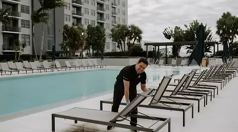 A smiling male cleaner adjusting a lounge chair on an outdoor swimming pool deck next to an apartment complex.