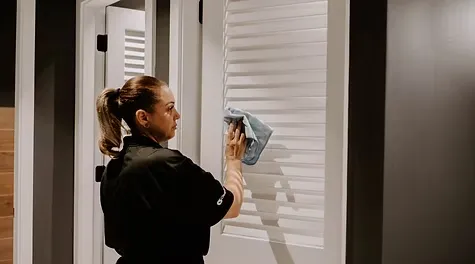A female cleaner wiping down white louvered door slats with a cloth.