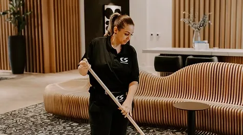 A female cleaner in a black uniform sweeping a patterned rug in a modern lounge area.