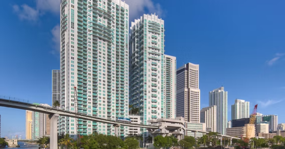 Modern high-rise skyscrapers with an elevated monorail track running past in the foreground under a blue sky.