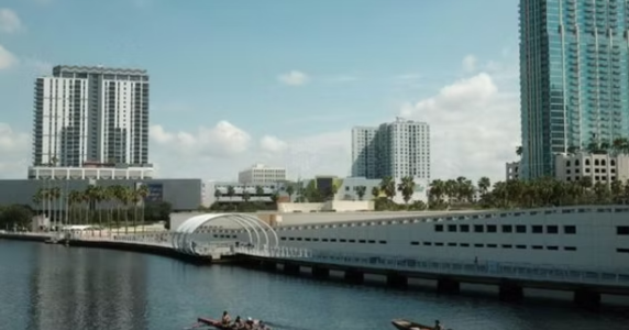 Modern city waterfront skyline featuring high-rise buildings, palm trees, a pedestrian boardwalk, and rowers on the water.