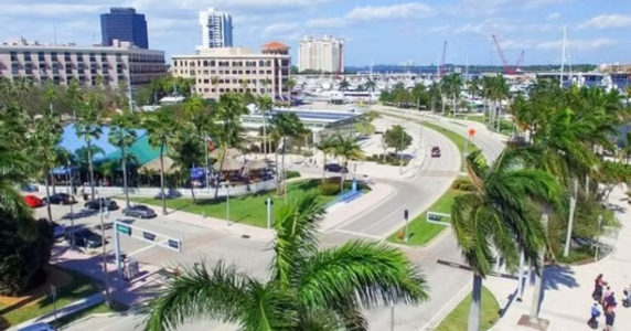 Aerial view of a coastal city street lined with palm trees, multi-story buildings, and a marina in the background.