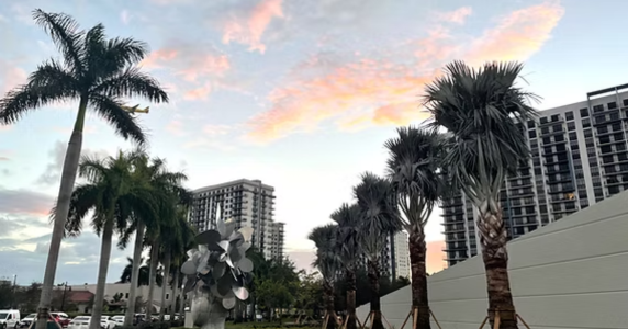 Modern high-rise residential or commercial buildings framed by palm trees and outdoor sculpture against a sunset sky.