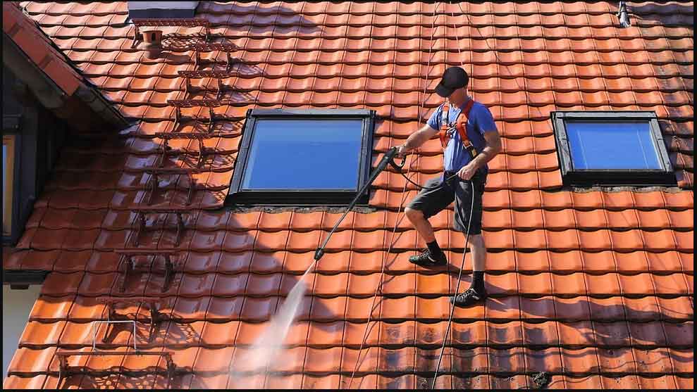 Worker wearing a safety harness pressure washing a terracotta tile roof near skylights.