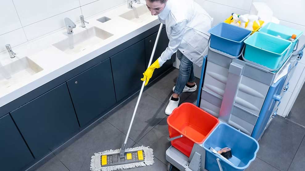 Janitor mopping a tiled commercial restroom floor next to a cleaning cart with buckets and supplies.