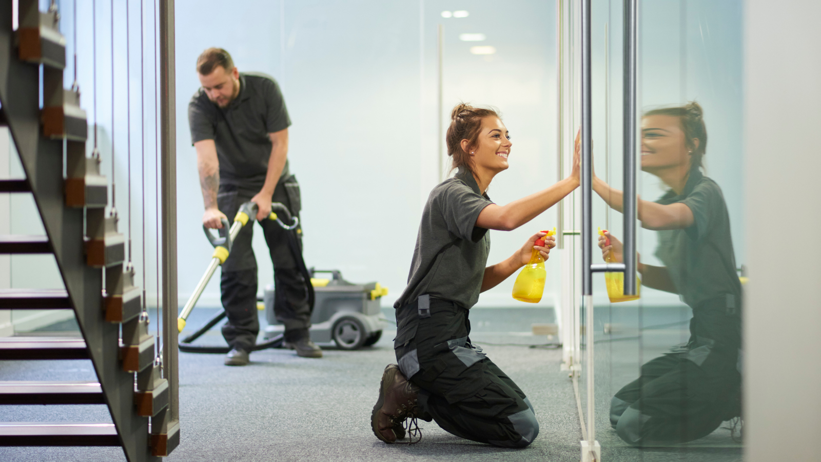 Two commercial cleaning staff working in an office, with one woman wiping down a glass door and a man vacuuming the carpet in the background.